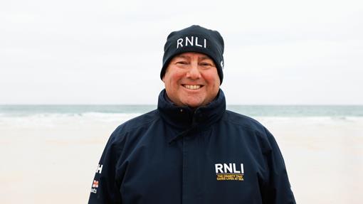 RNLI face-to-face fundraiser David, smiling in a blue RNLI jacket and hat, with Porthmeor Beach, St Ives, in the background